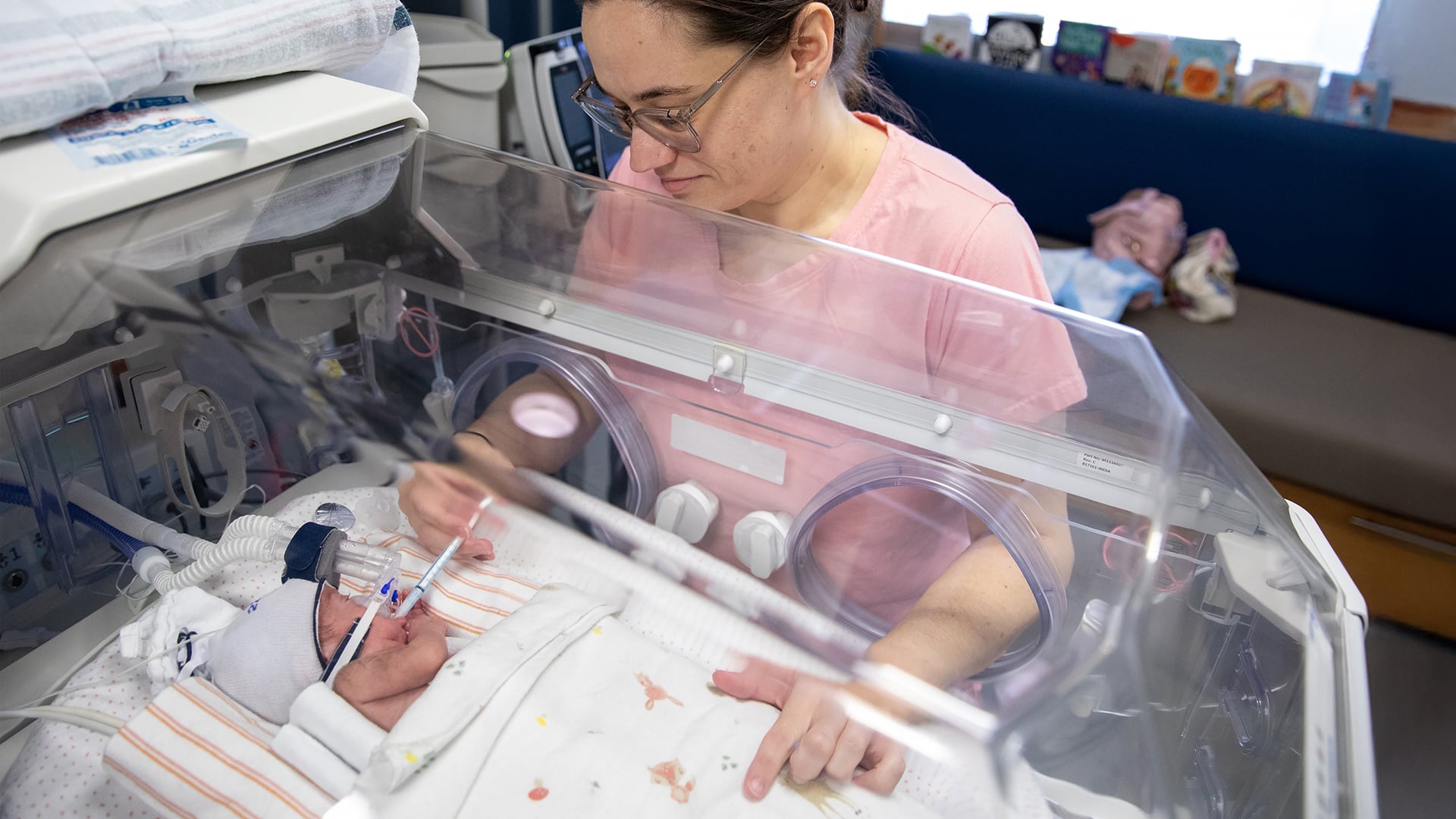 A woman in a pink t-shirt leans over an incubator in the NICU, feeding a premature baby donor milk through a syringe while the infant rests under soft blankets and breathing support.