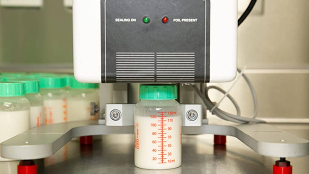 A filled milk bottle sits under a sealing machine, surrounded by additional bottles waiting on a rotating platform.