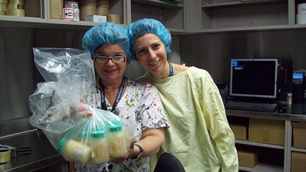Two staff members wearing hairnets smile while holding a large plastic bag filled with milk bottles inside a processing room.