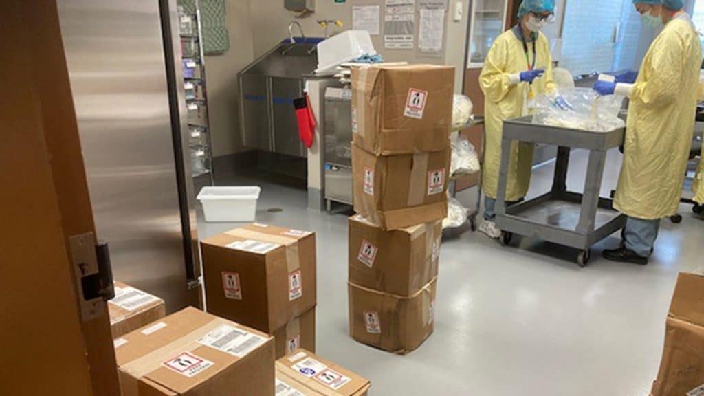 Staff in PPE process shipments in a receiving area filled with stacked frozen-milk delivery boxes and stainless steel workstations.