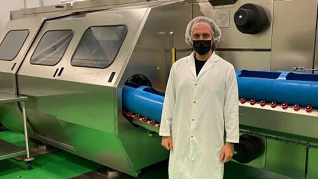 A researcher wearing a white lab coat, hair cover and face mask stands in front of large stainless steel processing equipment inside a laboratory or production facility.