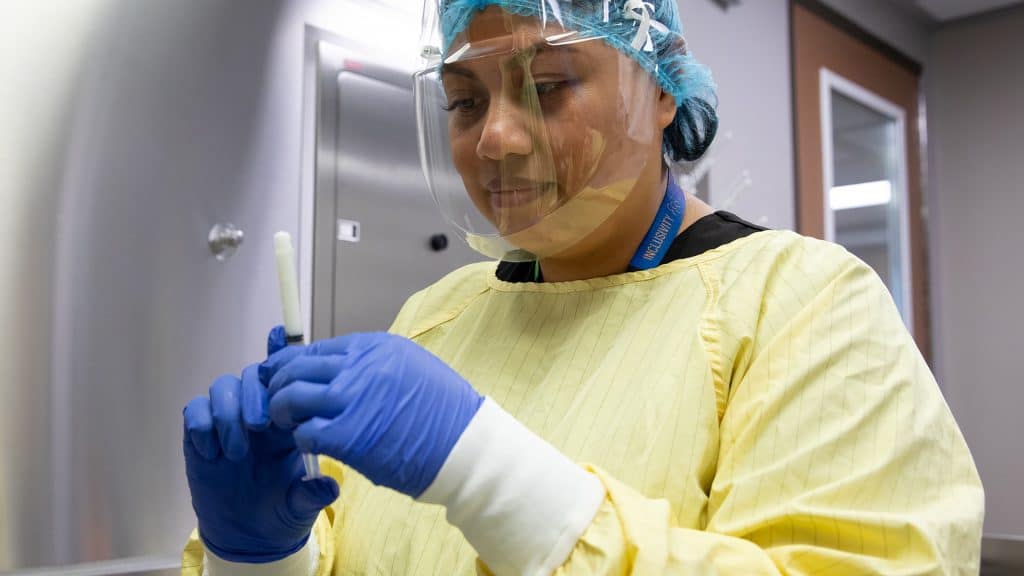 A diet technician wears protective gear, including gloves, a gown, a face shield and a hair cover, while preparing equipment in a clean processing room.