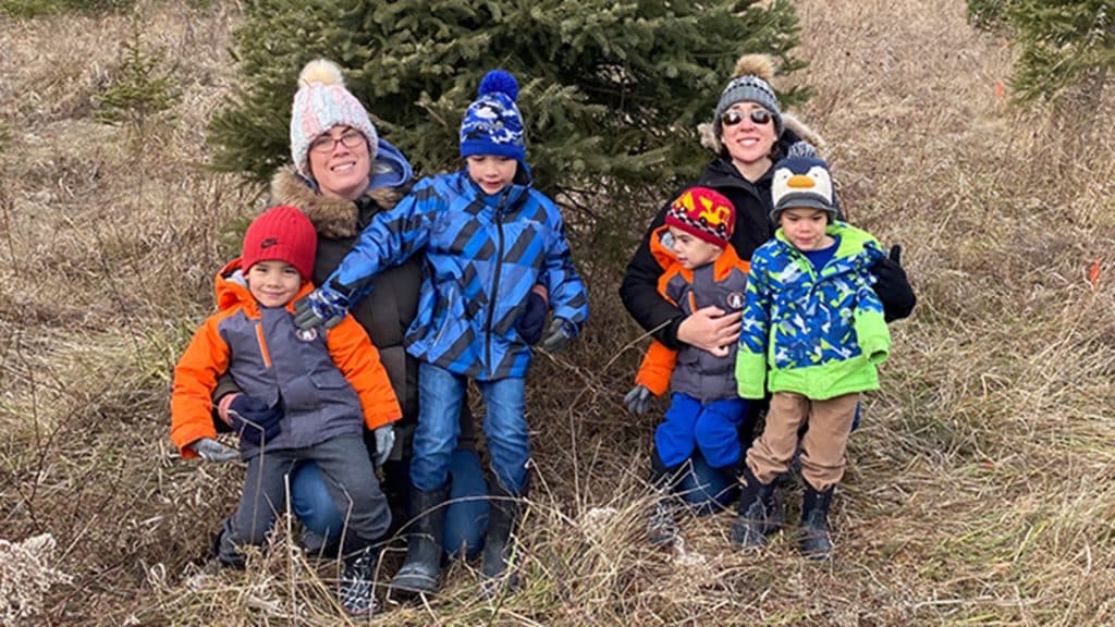 Two adults and four young children pose together outdoors in winter clothing, standing in front of evergreen trees with dried grass beneath them.