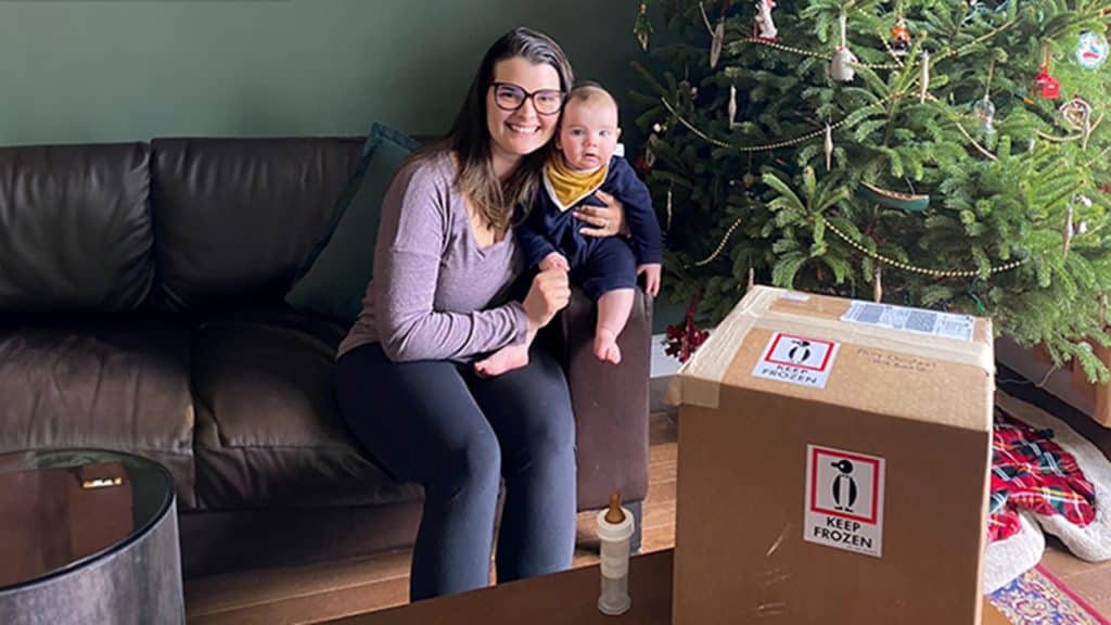 A milk donor named Amanda sits on a couch holding her baby beside a large shipping box labeled "Keep Frozen," with a decorated Christmas tree in the background.