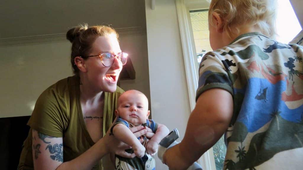 Milk donor Ambrosia smiles while holding her baby as an older child stands nearby, the family gathered in a bright room with natural light streaming in.