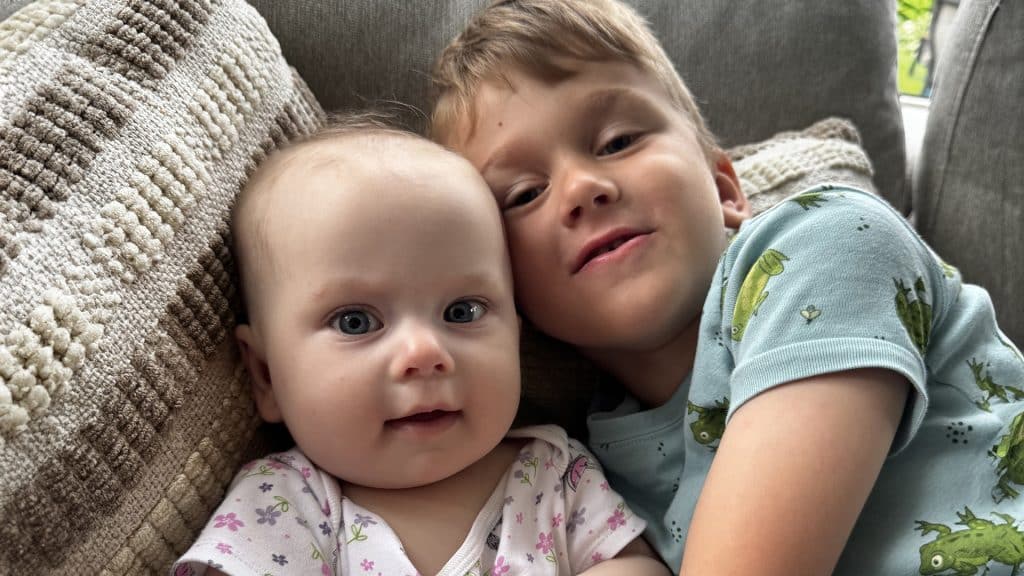 A baby and an older sibling cuddle together on the couch, smiling gently while resting on a couch.