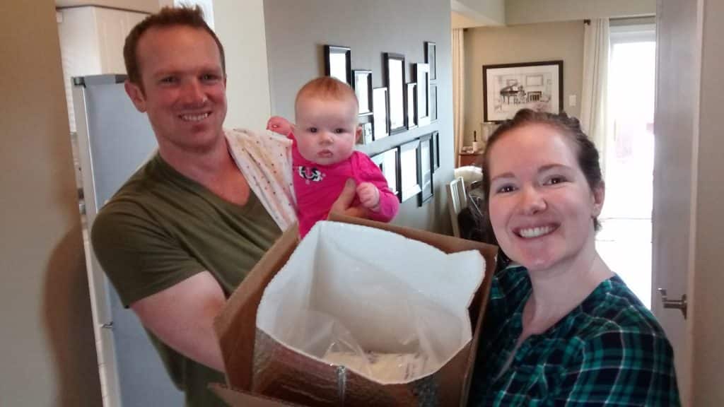 Milk donor Brooke stands beside her partner, who is holding their baby and a large insulated box filled with donated milk, all smiling inside their home.