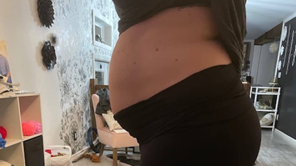 A pregnant donor shows a side view of her baby bump while standing in a nursery, with toys and decor visible in the background.