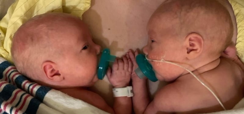 Two newborn twins lie facing each other on a blanket, each holding a pacifier and gently touching hands, with one baby supported by a small nasal breathing tube.