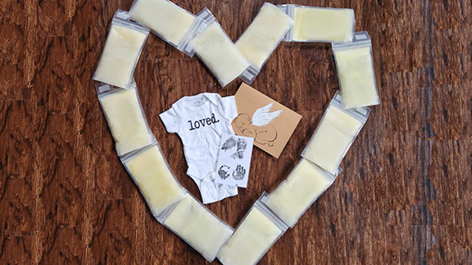 Donor milk bags are arranged in the shape of a heart around a baby onesie that reads "loved" and two small keepsakes, all placed on a wooden surface.