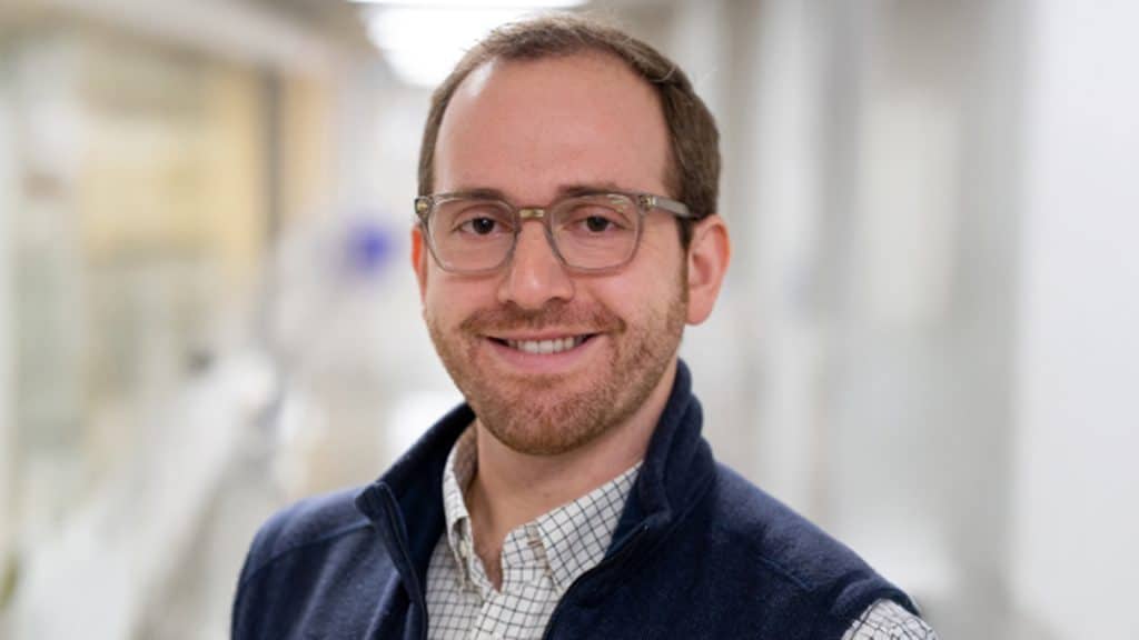 Michael Pitino, director of the Milk Bank, smiles in a professional headshot, wearing glasses, a collared shirt and a dark vest, with a softly blurred background behind him.