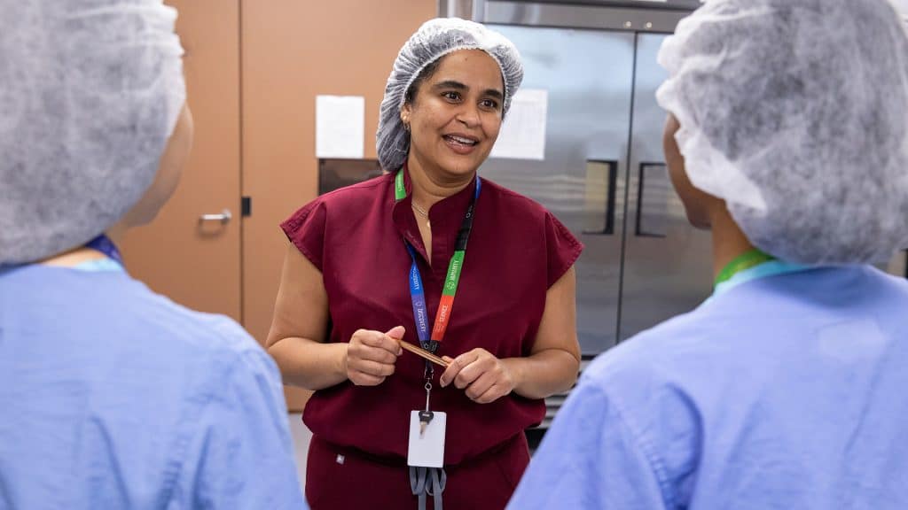 Production supervisor Sheena Ragoo, wearing a hairnet and red scrubs, speaks to two staff members in blue scrubs and hairnets inside a milk bank processing area.