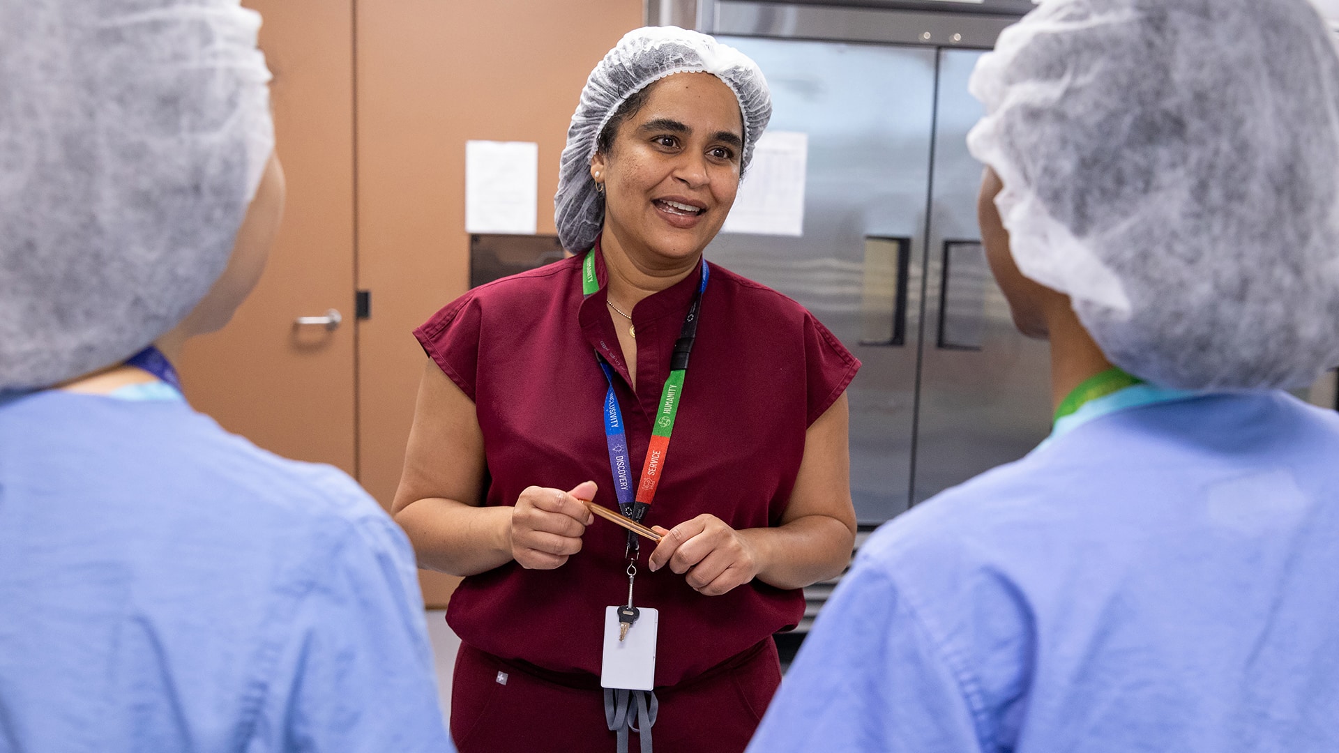 Production supervisor Sheena Ragoo, wearing a hairnet and red scrubs, speaks to two staff members in blue scrubs and hairnets inside a milk bank processing area.