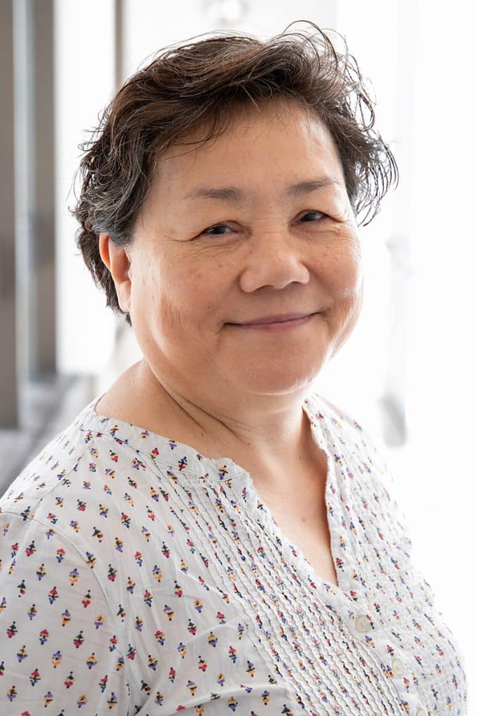 Carol Cheng smiles in a professional headshot wearing a light patterned blouse and standing in front of a bright, softly blurred background