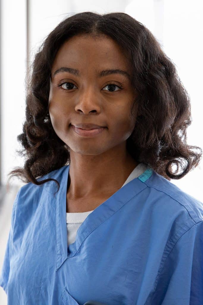 Chelsea Denton poses for a professional headshot, wearing blue scrubs and standing in front of a bright neutral background