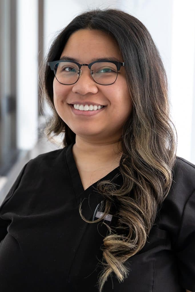 Elisa Andaya smiles in a professional headshot, wearing black scrubs and standing in front of a softly lit indoor background