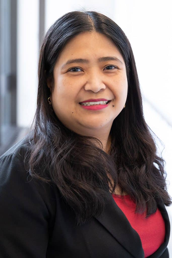 Holland Santos smiles in a professional headshot, wearing a pink top with a black blazer and standing in front of a softly lit indoor background
