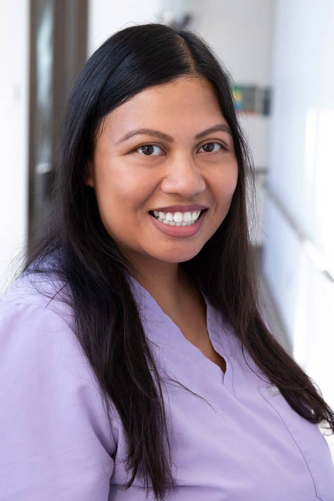 Jessica Atan smiles in a professional headshot, wearing light purple scrubs and standing in front of a softly lit indoor background