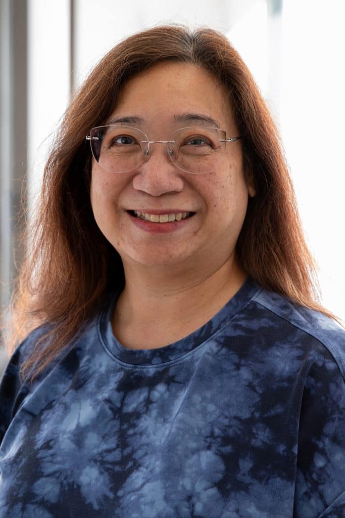 Karen Canals smiles in a professional headshot, wearing a blue patterned top and standing in front of a softly lit indoor background