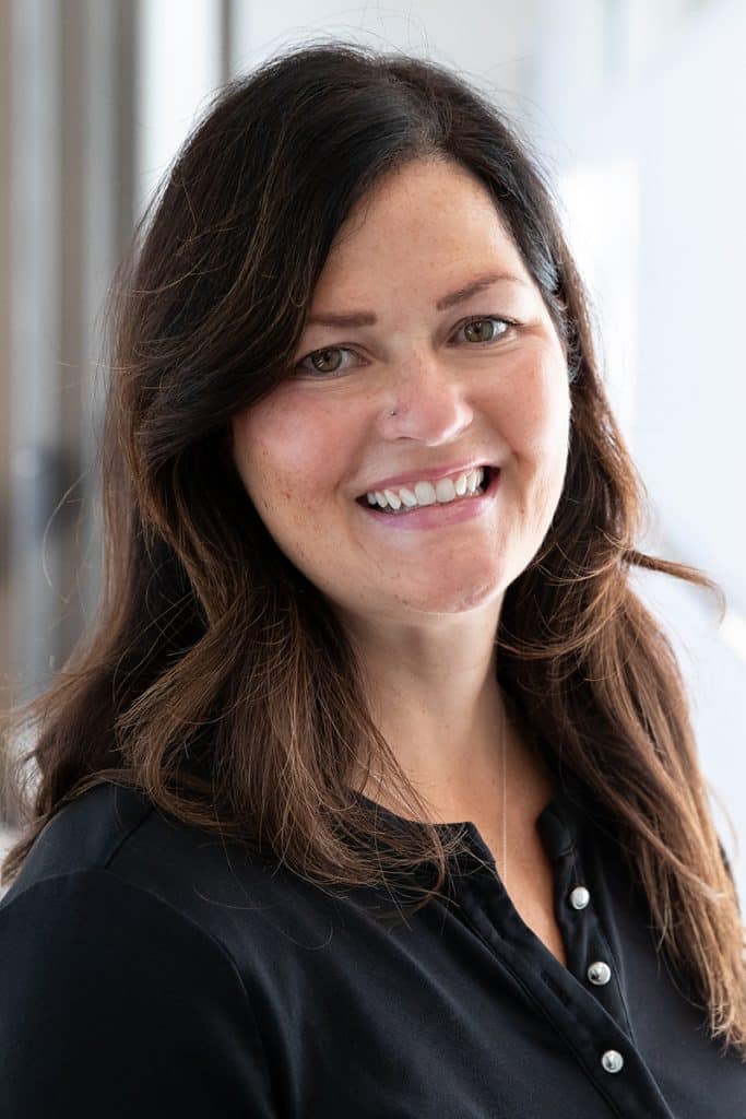 Natalie Shepherd smiles in a professional headshot, wearing a black top with buttons and standing in front of a softly lit neutral background