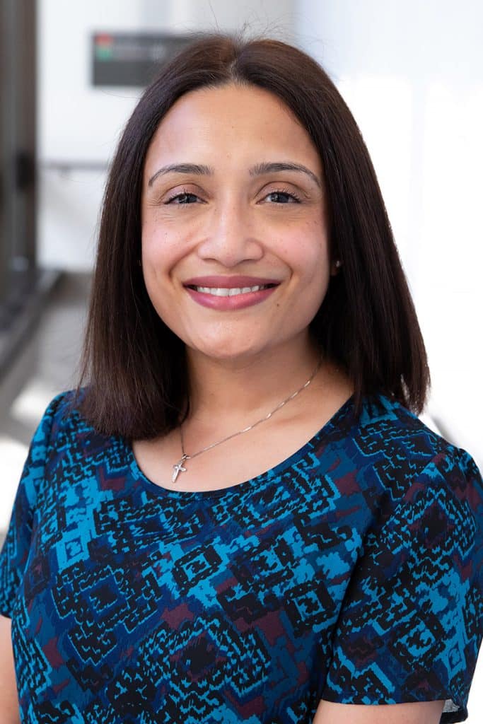 Sandra Fernandes smiles in a professional headshot, wearing a blue patterned top and standing in front of a softly lit indoor background