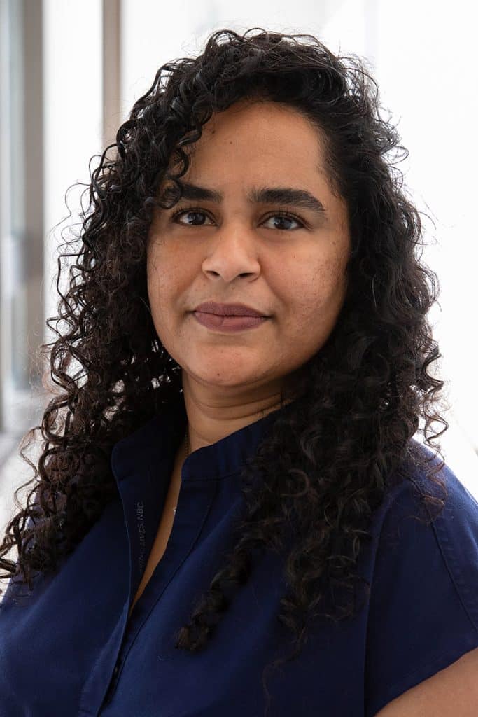 Sheena Ragoo poses for a professional headshot, wearing a navy top and standing in front of a bright, neutral background with soft lighting