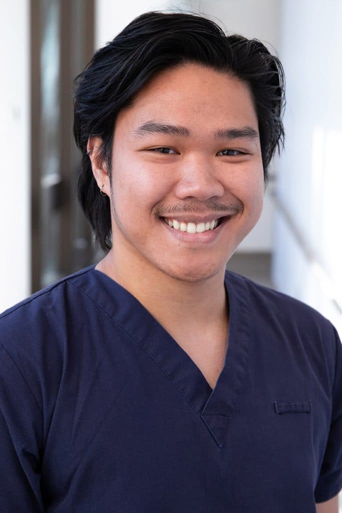 Tristan Ducay smiles in headshot, wearing navy scrubs and standing in front of a softly blurred indoor background